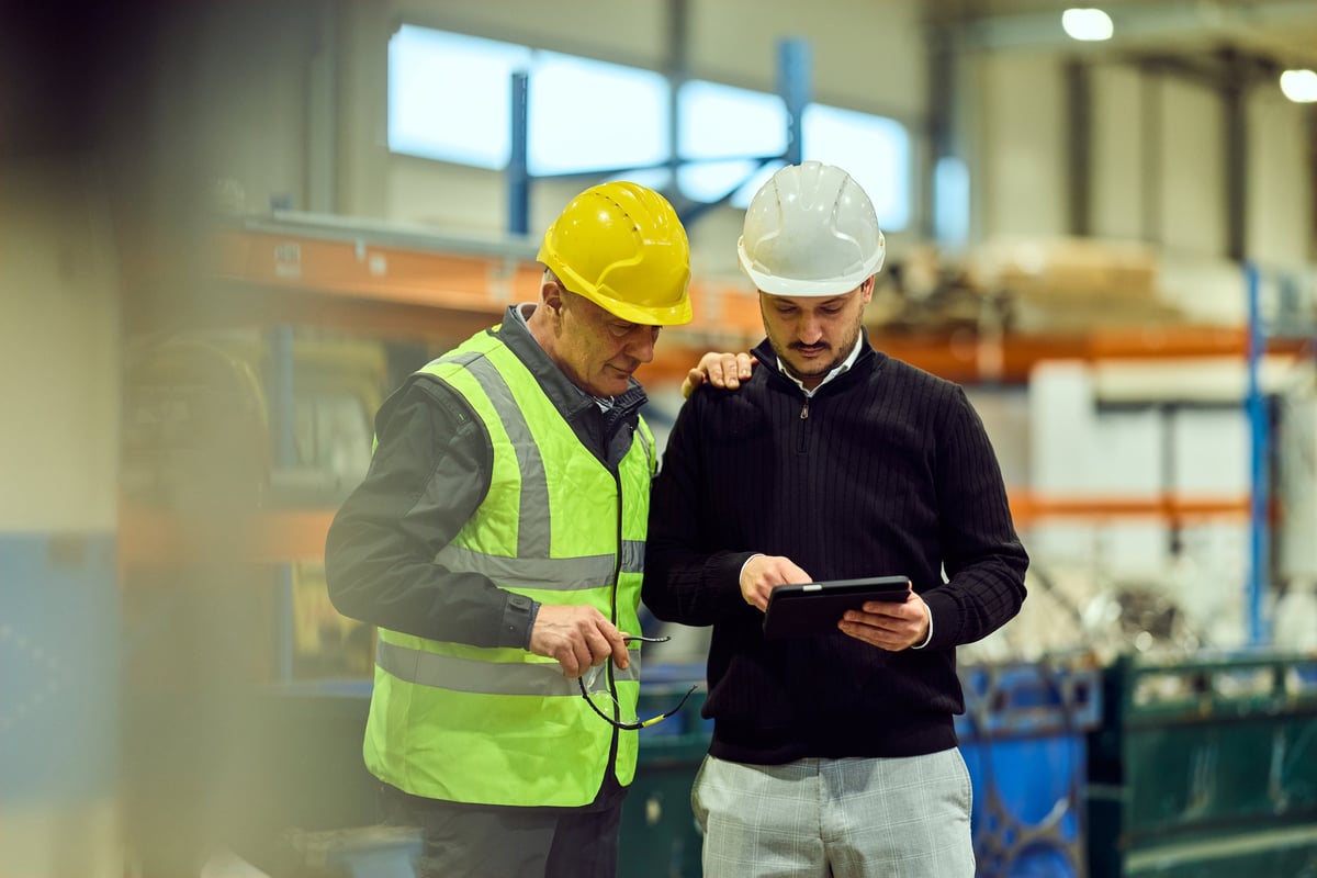 Two technicians in hard hats and high-visibility vests reviewing production data on a tablet inside an industrial materials facility