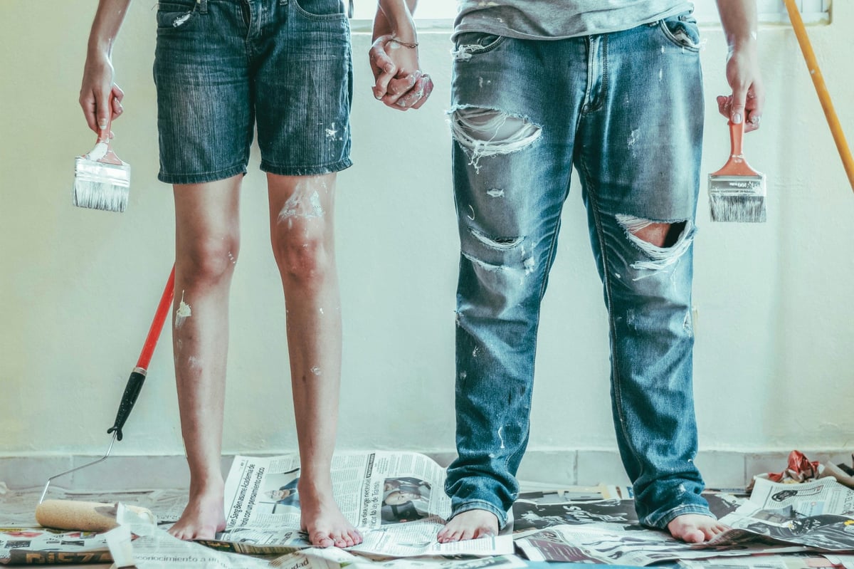 Two people holding paintbrushes and hands while decorating a room, with a roller and newspapers on the floor